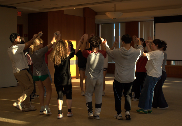 A group of students raises joined hands as they dance in a circle in the middle of a large room.
